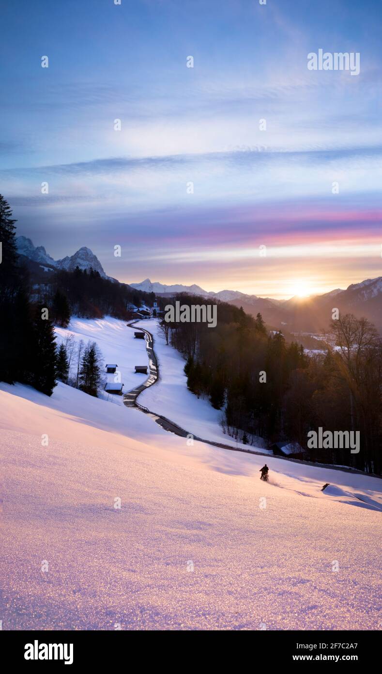 Beautiful pink sunset in the mountains with leading line road towards church and motorbiker having fun in fresh snow Stock Photo