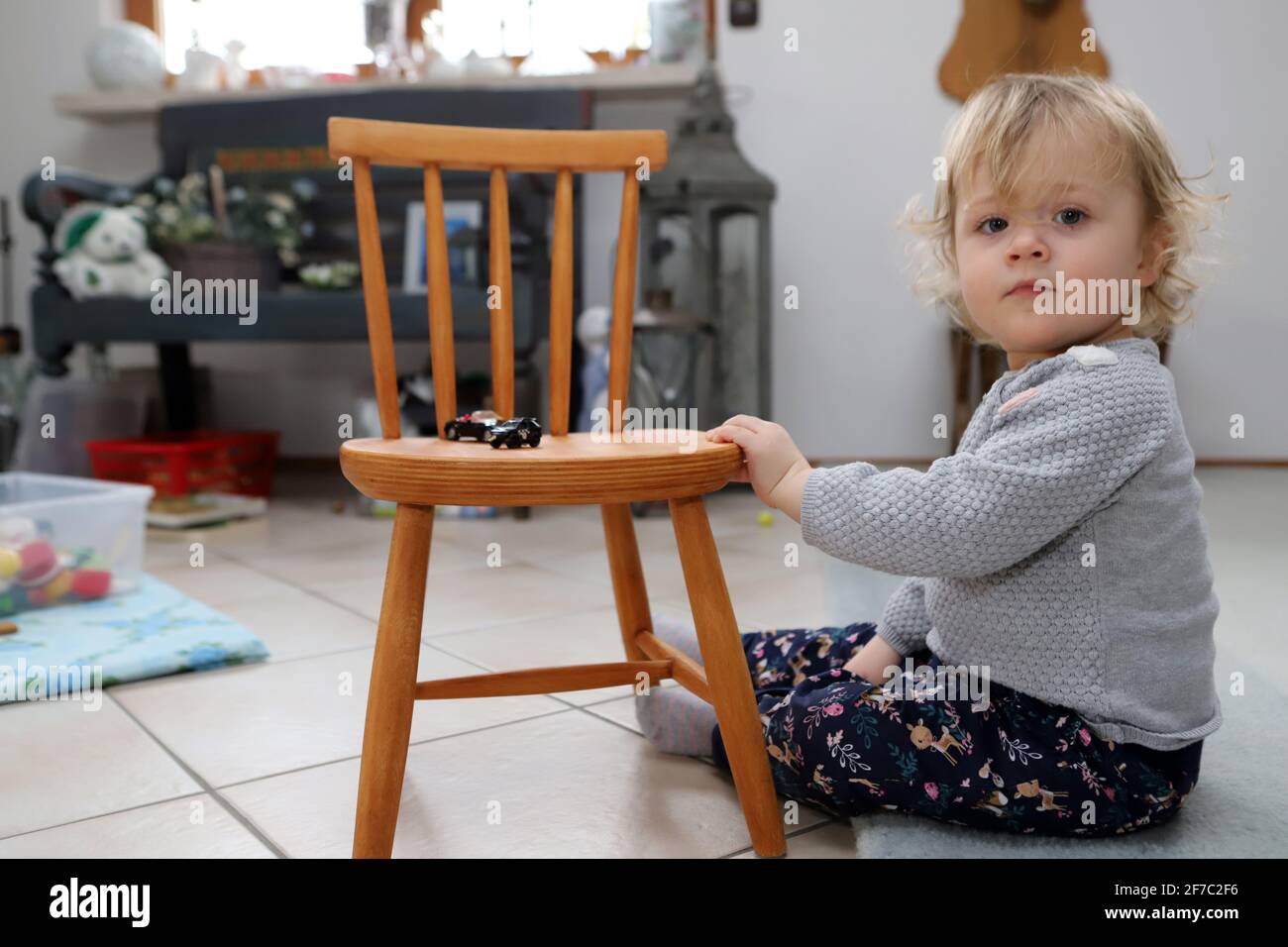 kleines Mädchen sitzt im Wohnzimmer auf dem Boden neben einem Kinderstuhl aus Holz -  little girl sits on the floor next to wooden child chair in livi Stock Photo