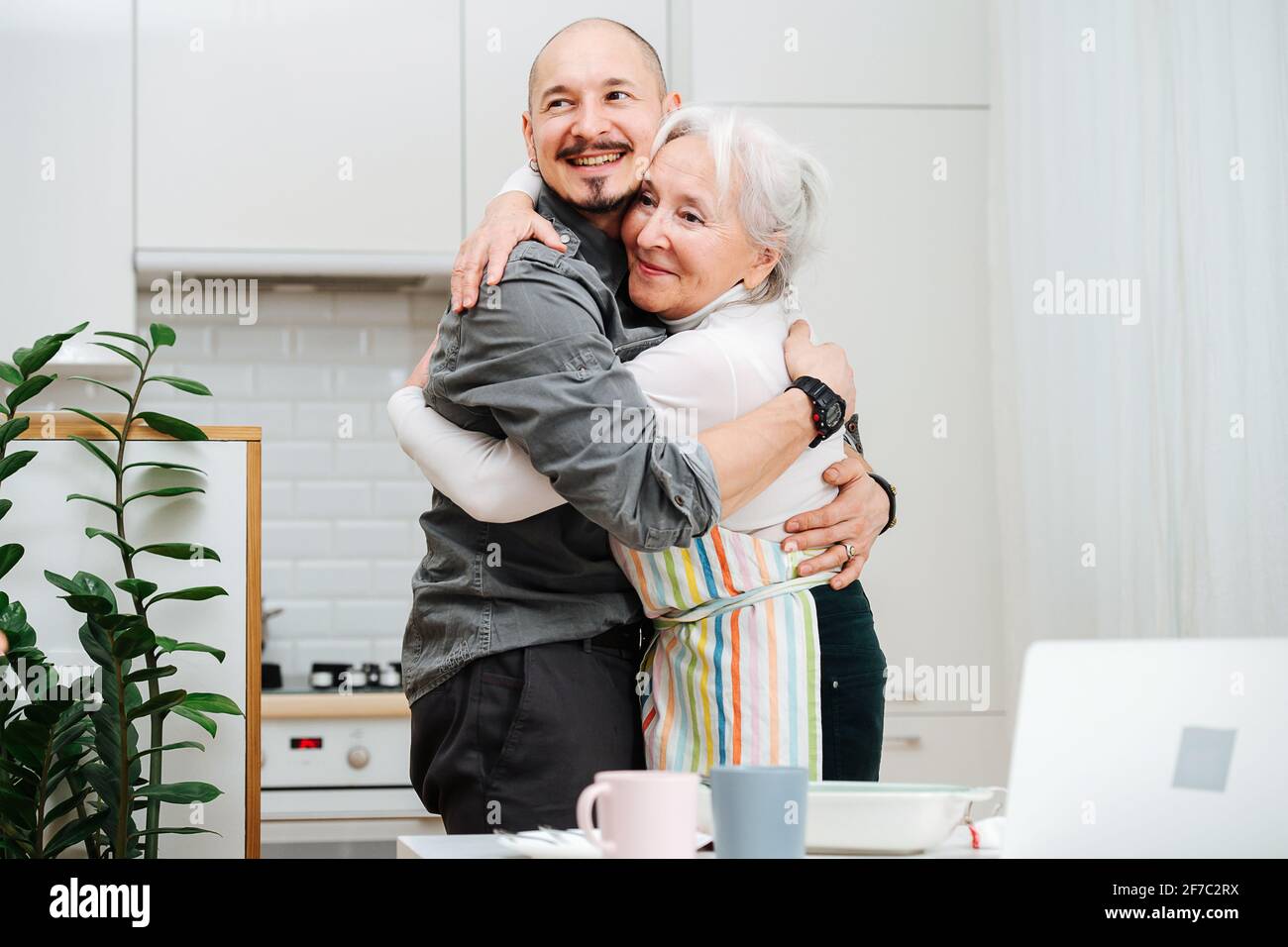 Smiling granny hugging her son like they didn't see each other for a long time. She wears apron, they stand in the kitchen. He is a lot taller than he Stock Photo