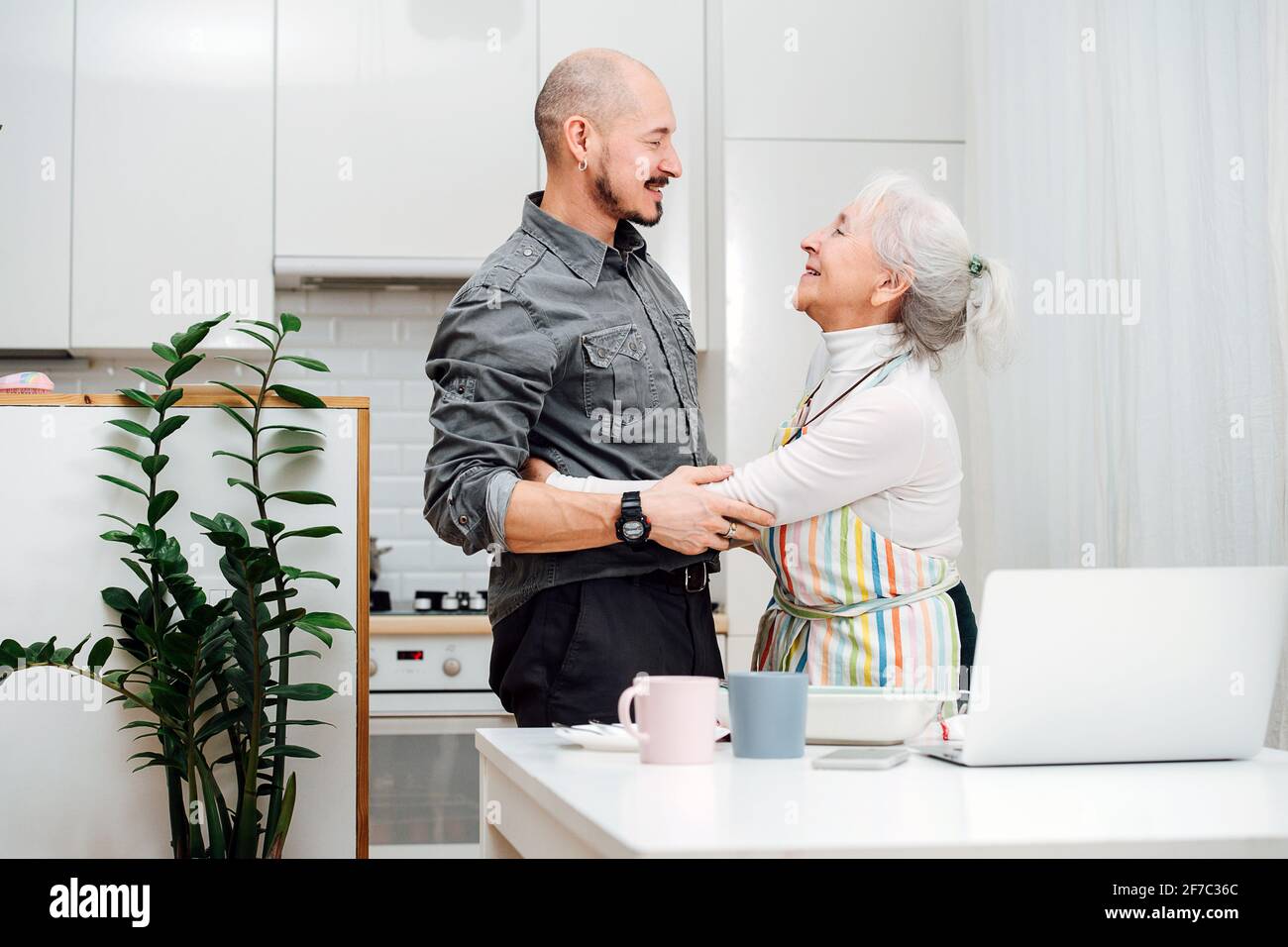 Happy granny and her son holding each others arms, greeting each other like they didn't see each other for a long time. She wears apron, they stand in Stock Photo