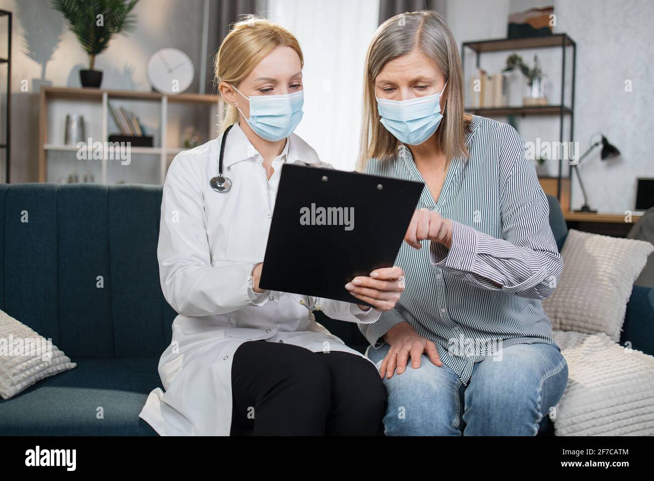 Mature woman doctor in white coat and stethoscope on shoulders talking to senior lady patient, sitting on sofa, showing treatment plan, medication list, explaining prescription and diagnosis Stock Photo