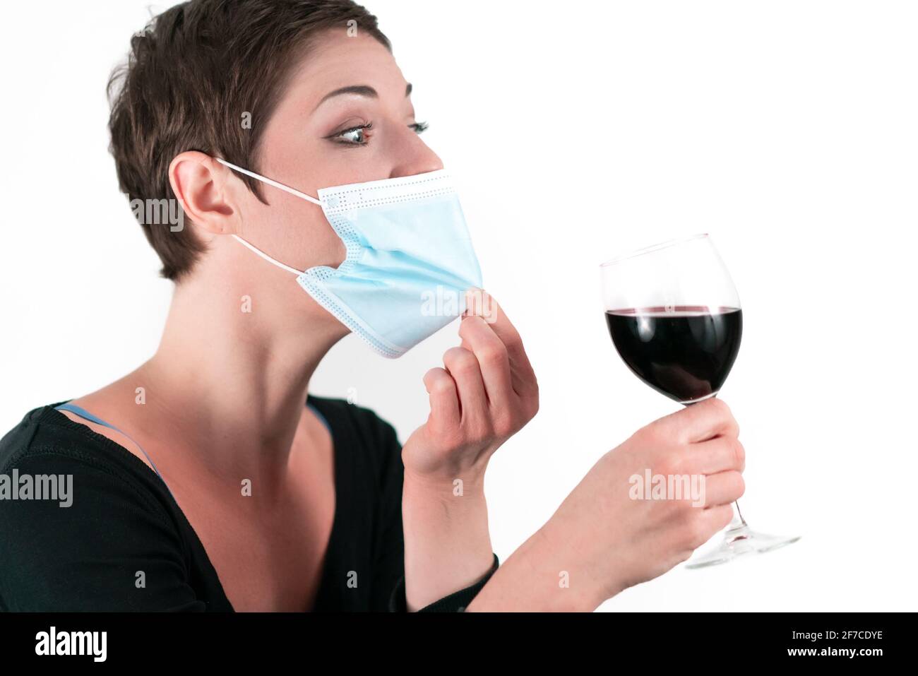 Beautiful woman with short hair wearing a face mask trying to find a way  how to drink from red wine from a glass. Life in quarantine. Dealing with  cor Stock Photo -