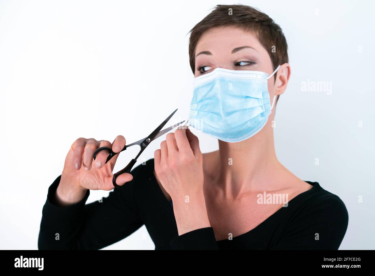 Beautiful woman with short hair removing her face mask with scissors. Life in quarantine. Dealing with coronavirus restrictions. Life during pandemic. Stock Photo