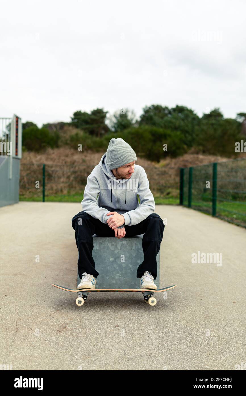 Portrait of a young adult male with a skateboard sitting on a box at a local skatepark. Skateboarding healthy lifestyle concept Stock Photo