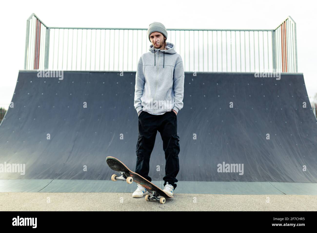 Portrait of a young adult male with a skateboard standing in front of a large ramp at a local skatepark. Skateboarding healthy lifestyle concept Stock Photo