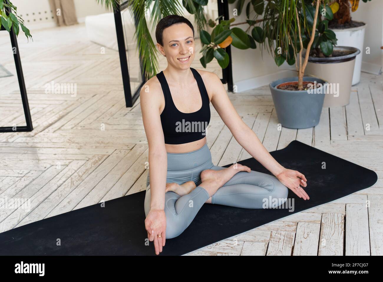 Young woman meditating indoors. pose Young woman smiling for the camera, sitting in the lotus position. Relaxation and meditation at home among the pl Stock Photo
