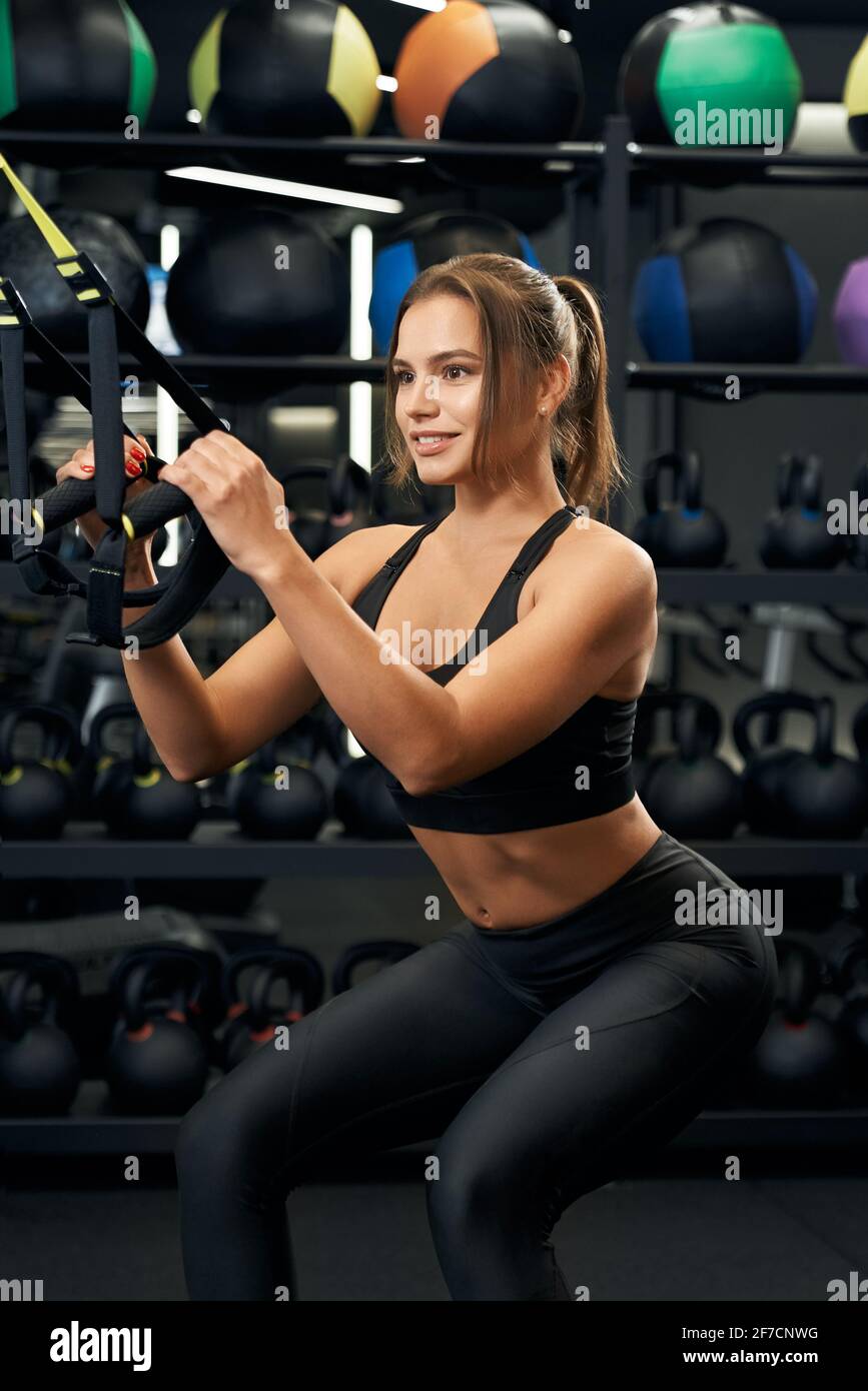 Close up of smiling sporty woman with beautiful body doing exercise and squatting with trx system. Concept of workout with special sports equipments in modern gym. Stock Photo