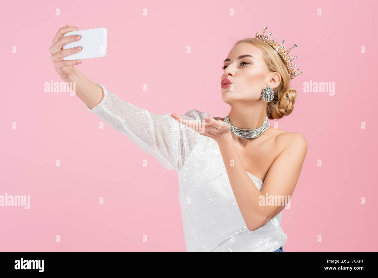 blonde woman in luxury crown taking selfie on smartphone and sending air kiss isolated on pink Stock Photo