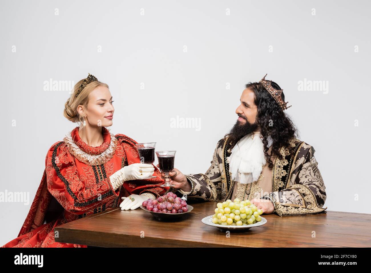 interracial historical couple in crowns clinking glasses of red wine while sitting at table with grapes isolated on white Stock Photo