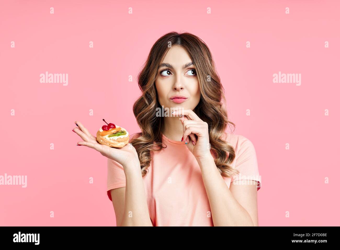 Dieting concept. Confused pretty woman looking up holding pastry cake in hand on pink background. Weight Loss Stock Photo