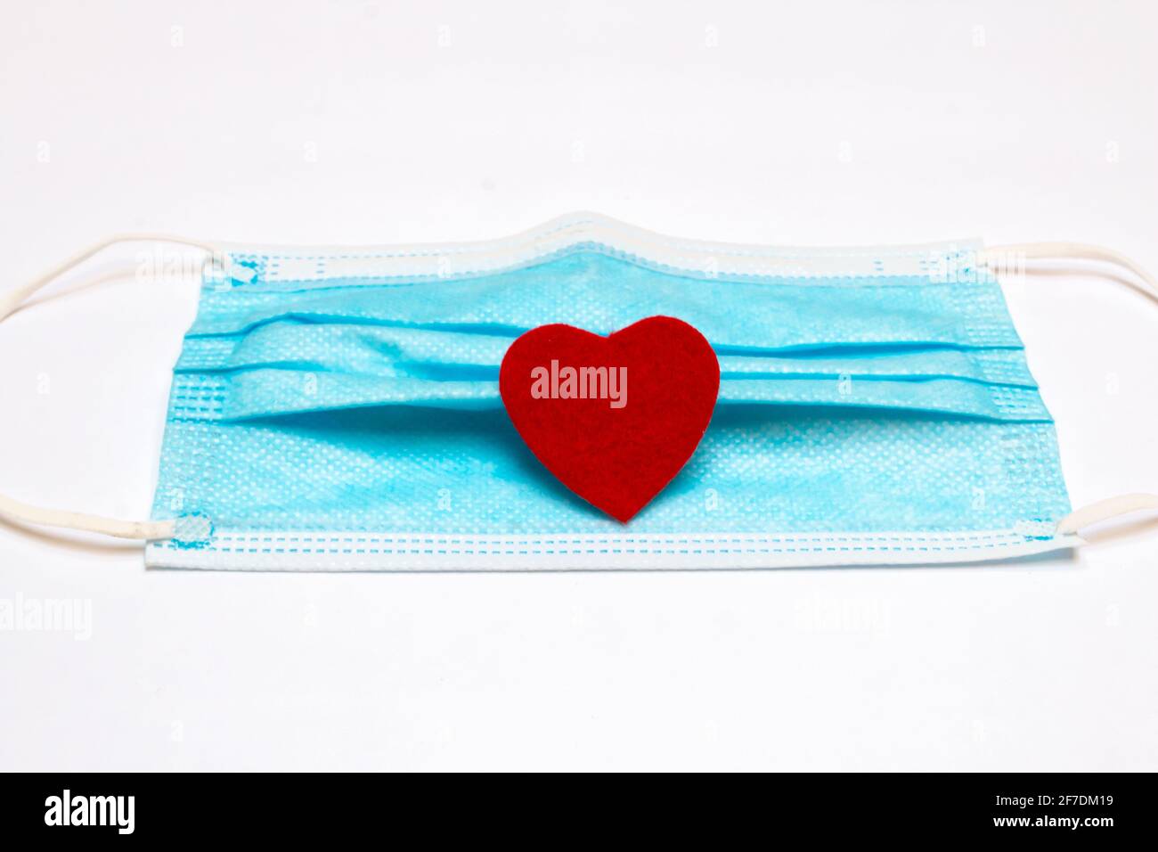 A blue medical mask with a small red felt heart on its folds, February 2021, COVID-19 Valentine's Day-themed. Stock Photo