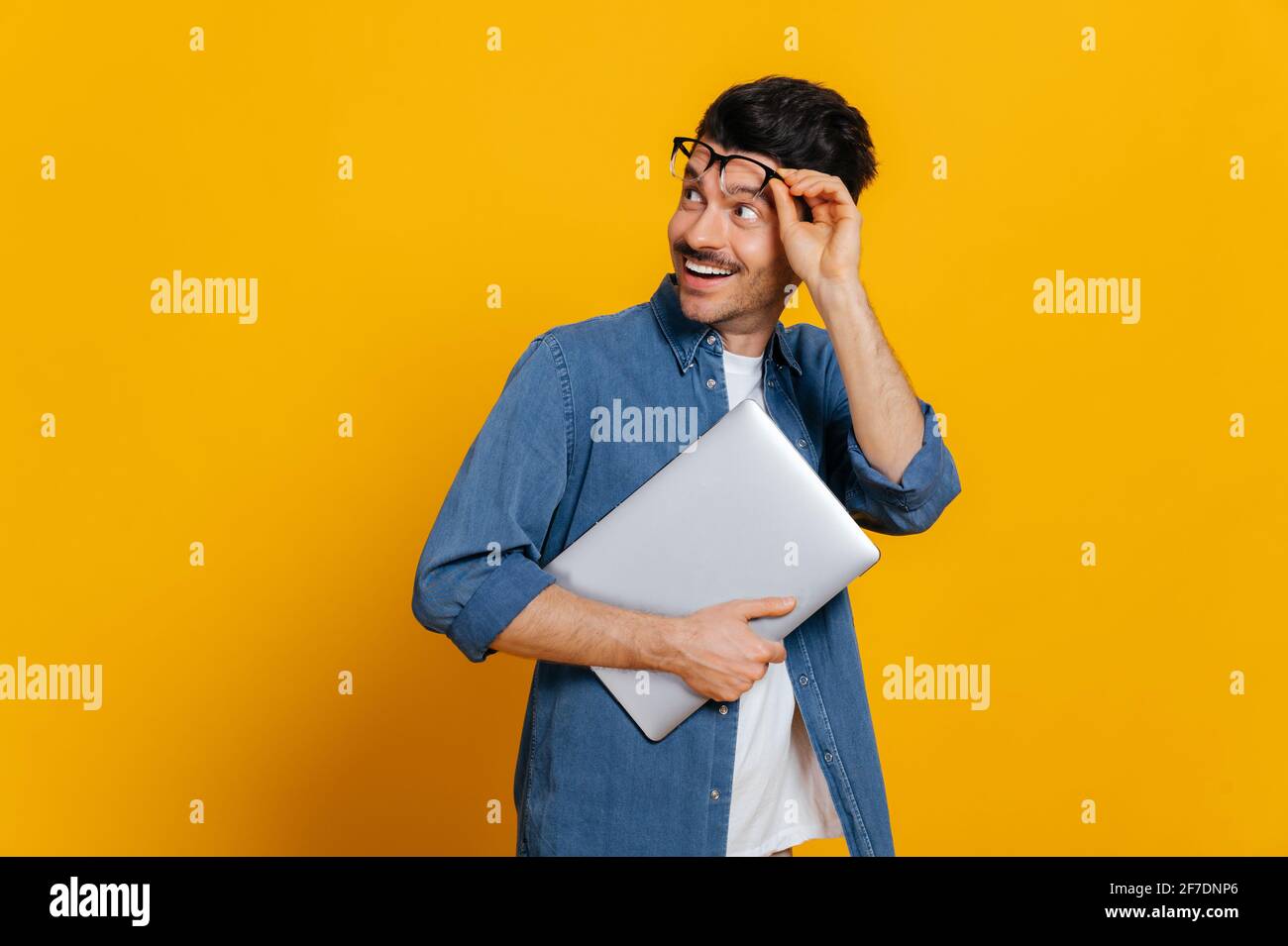 Amazed excited charismatic guy with glasses and stylishly dressed, holds a laptop at hand, looks surprised towards empty space taking off glasses, smiling, isolated orange background, copy space Stock Photo