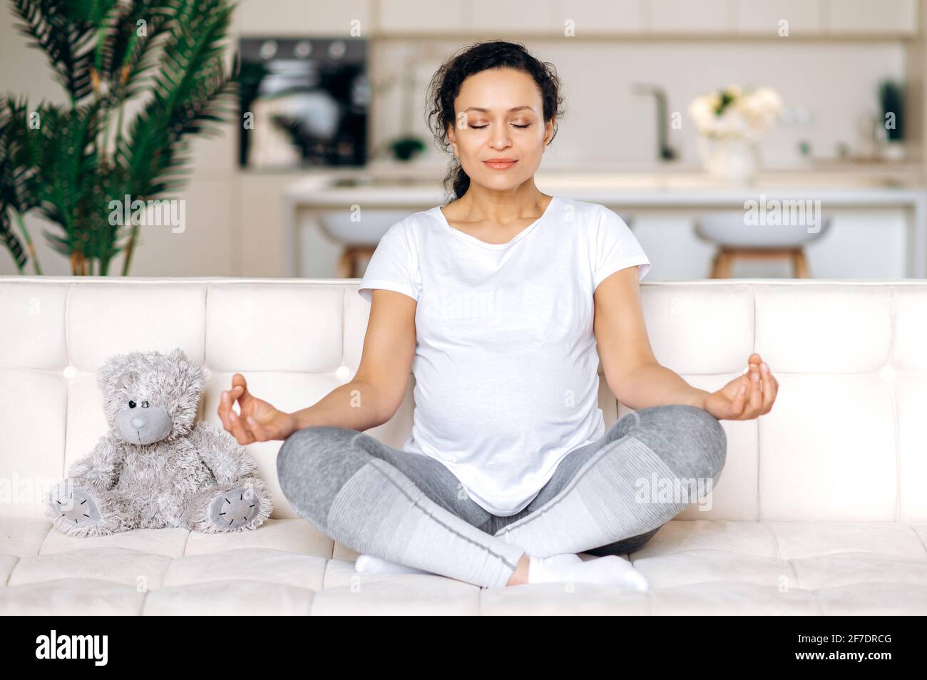 Attractive contented, beautiful future mom, brunette, mixed race, sitting on sofa at home in living room, in lotus position with closed eyes relaxes and meditates, focusing on exercises Stock Photo