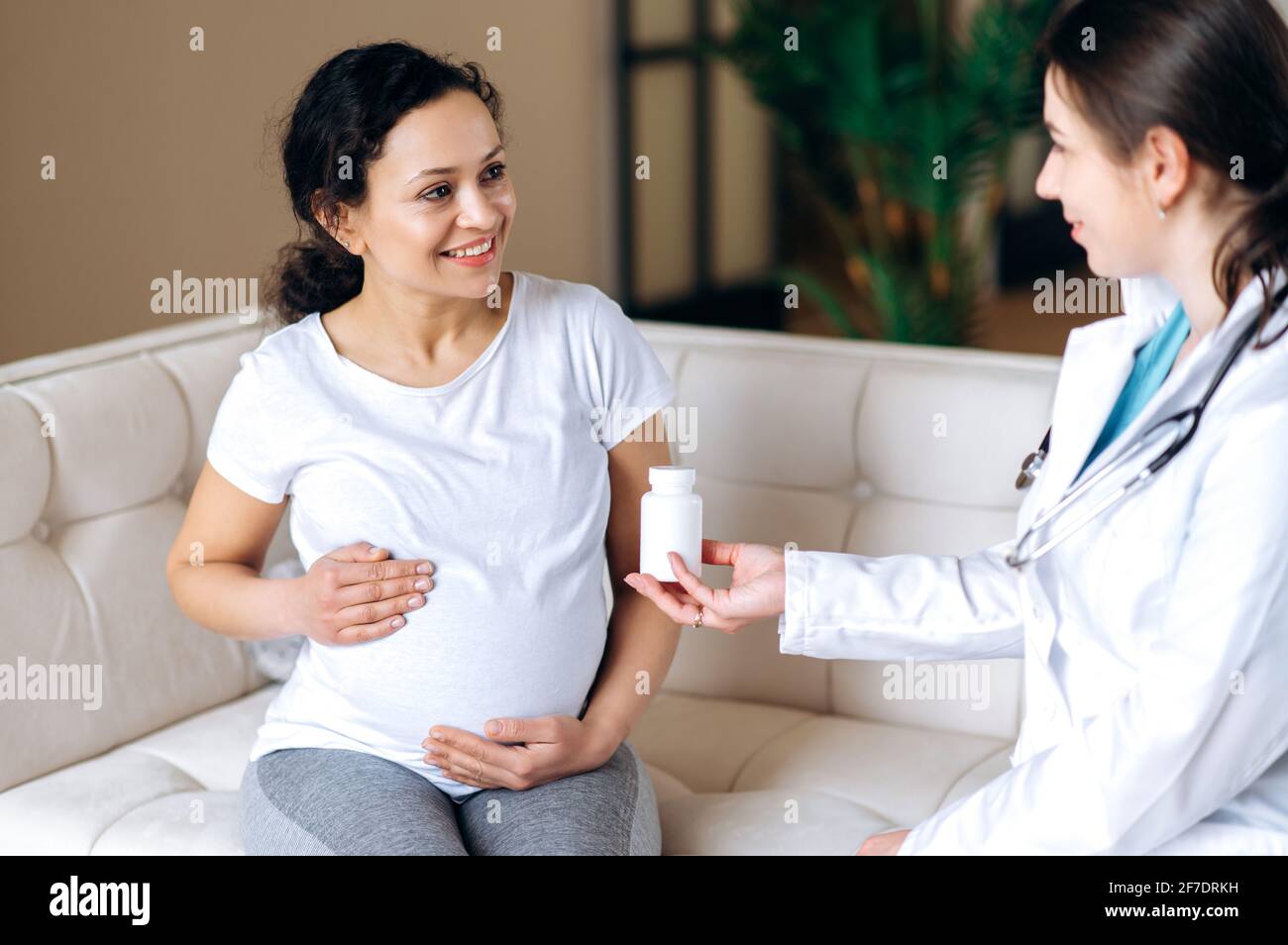 The doctor prescribes vitamins for a pregnant mixed race patient, sitting on the couch at home or in the hospital, the expectant mother stroking her belly. Healthy pregnancy concept Stock Photo