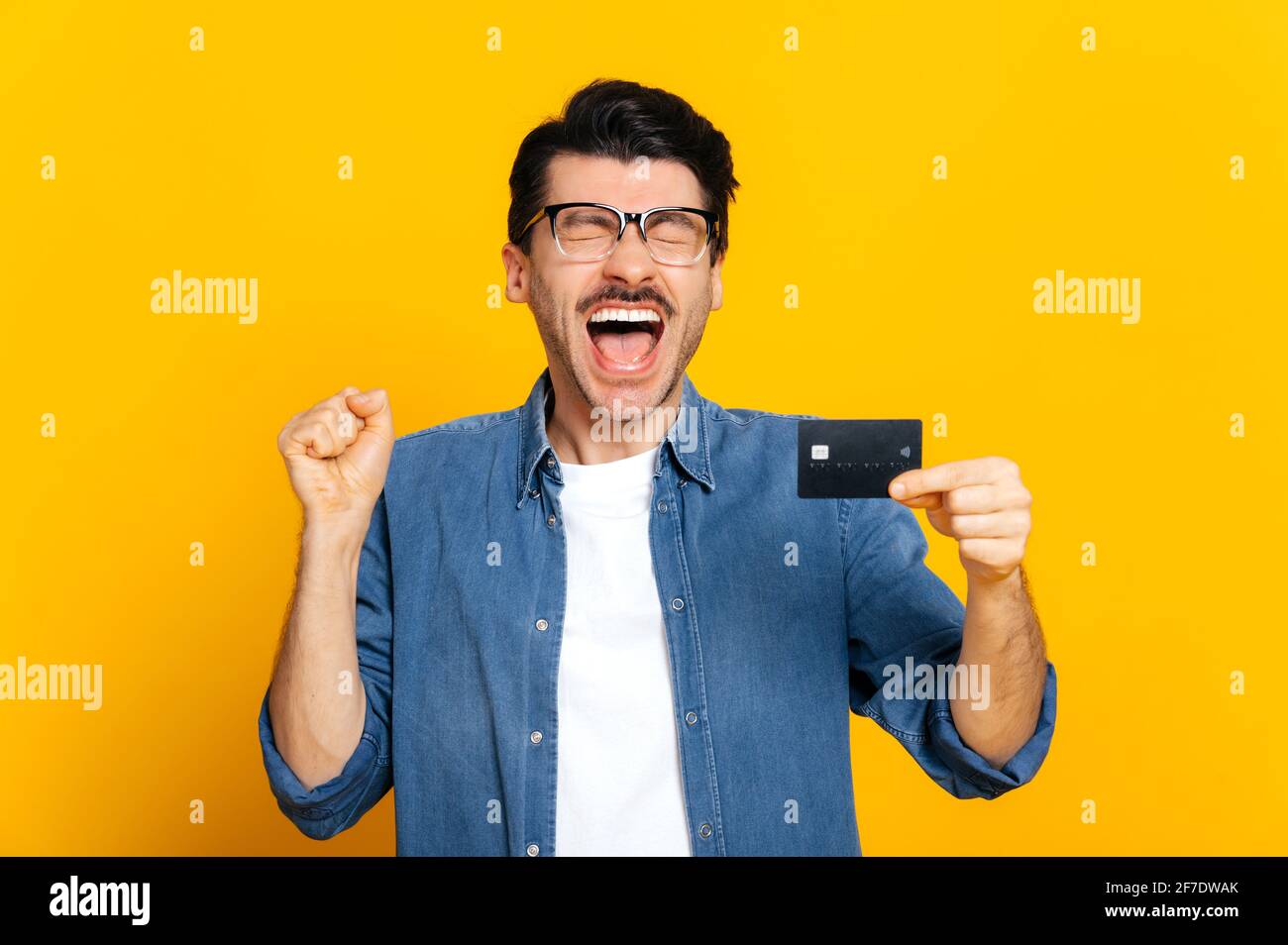 Happy cheerful caucasian unshaven stylish guy holding credit card in hand, shouting joyfully, standing on isolated orange background, finance and banking concept Stock Photo