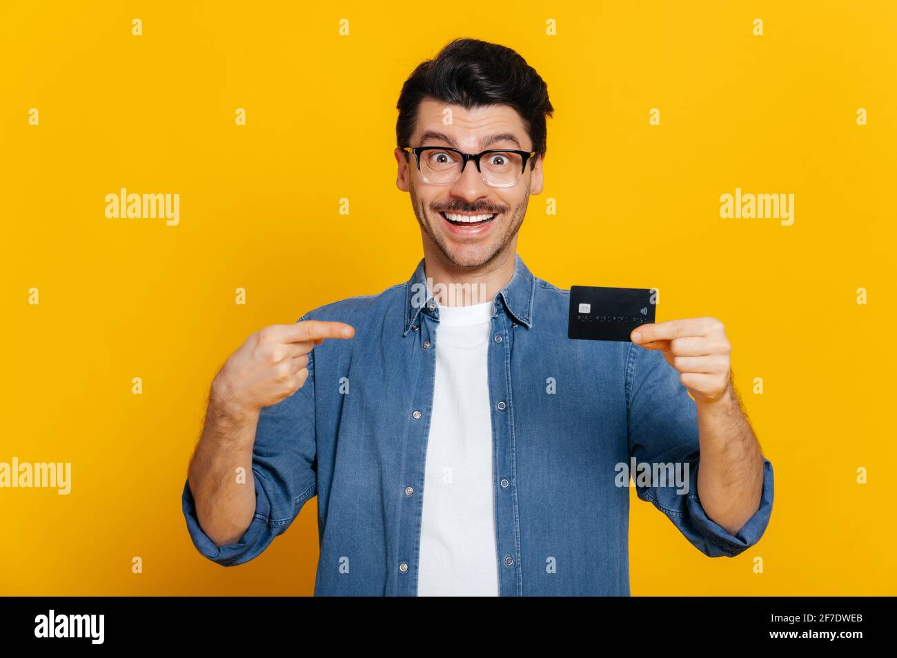 Amazed excited stylish handsome caucasian guy with glasses, holds a credit card in hand and points finger at it, looks happily at camera, smiling, stand on isolated orange background,e-banking concept Stock Photo