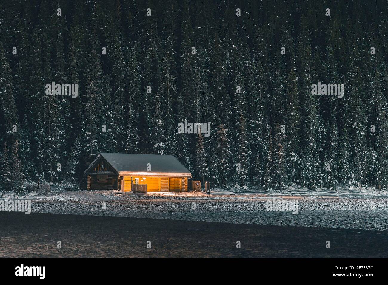 Small cabin on the banks of frozen Lake Louise during late winter night with snow capped trees around and in the background. Stock Photo