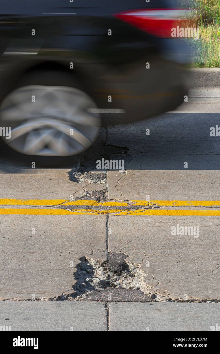Old worn concrete road highway pavement in disrepair showing cracks, patches, repairs and chuck holes as auto passes by, Castle Rock Colorado USA. Stock Photo