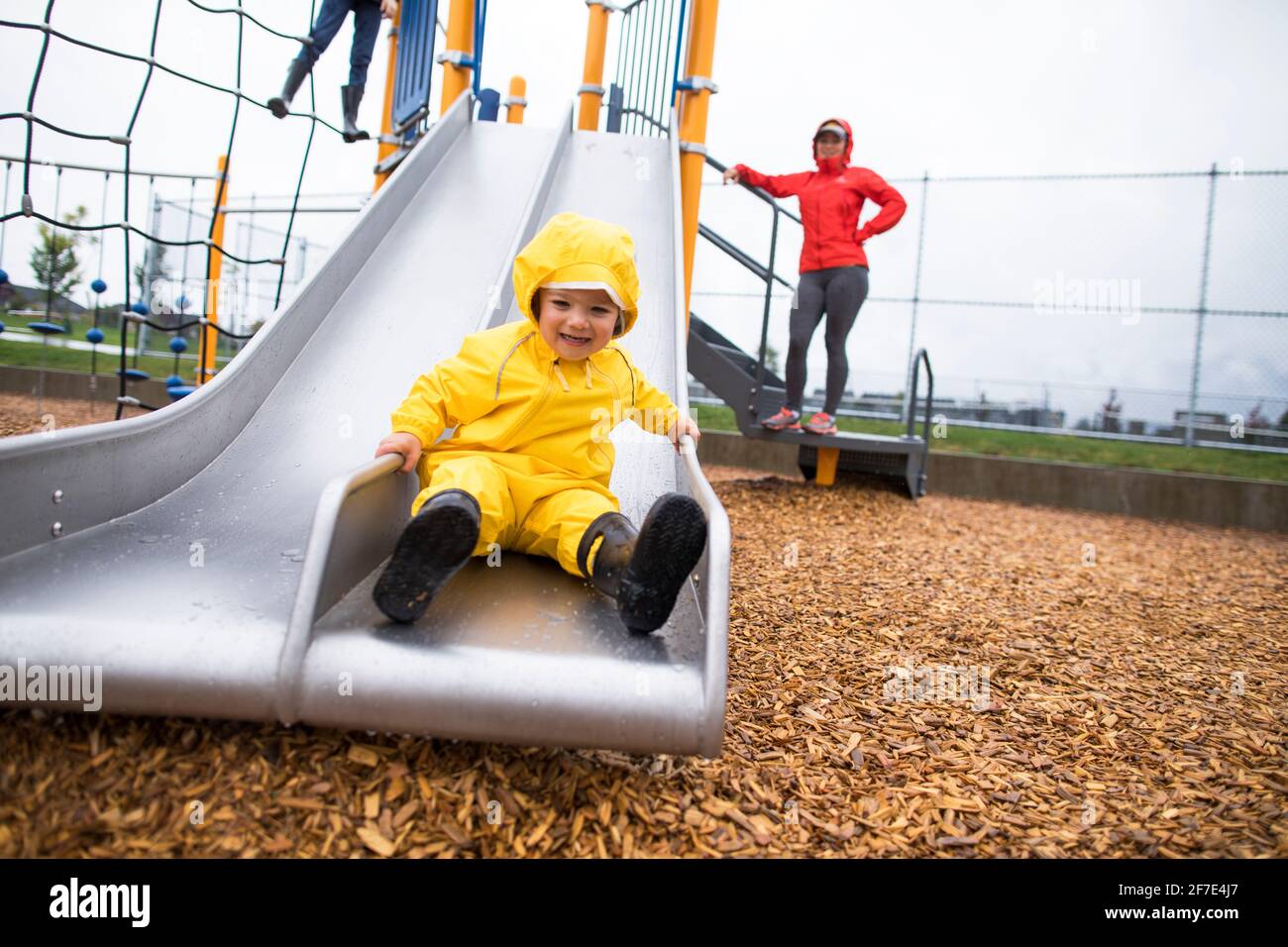 Happy boy sliding on slide at local park, mother watching. Stock Photo