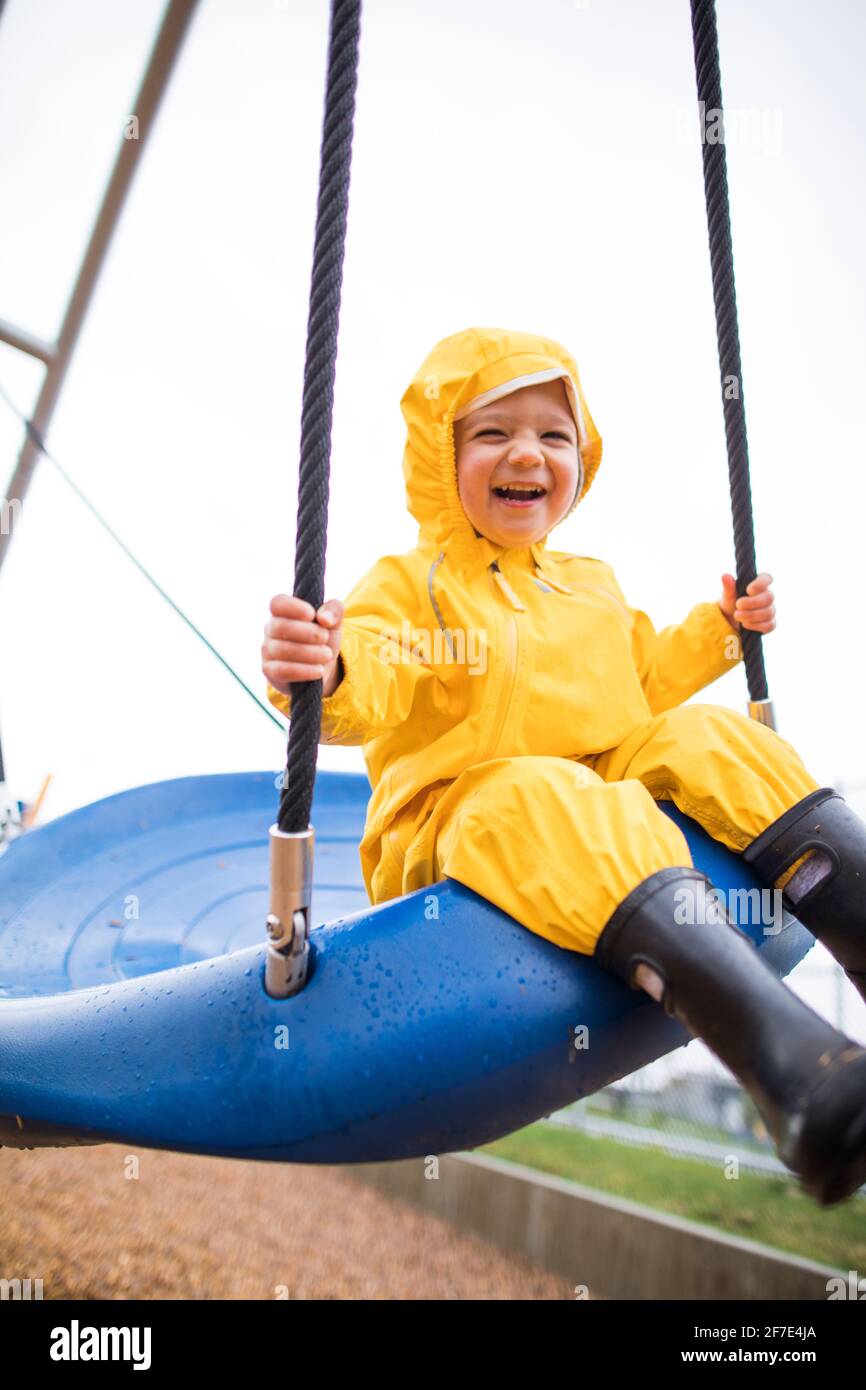 Happy toddler boy rides on swing at the park on a wet day Stock Photo