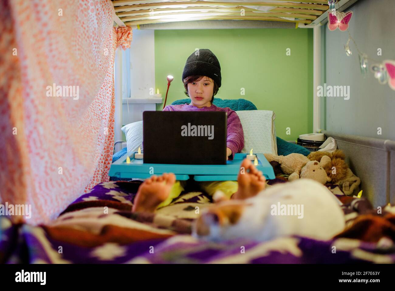 A boy sits in bed with knit hat doing schoolwork on computer with cat Stock Photo