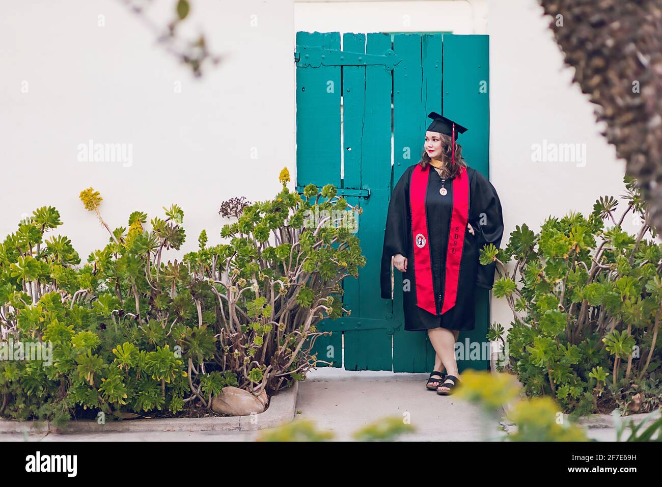 Woman wearing a graduation gown/cap standing in front of blue gate. Stock Photo