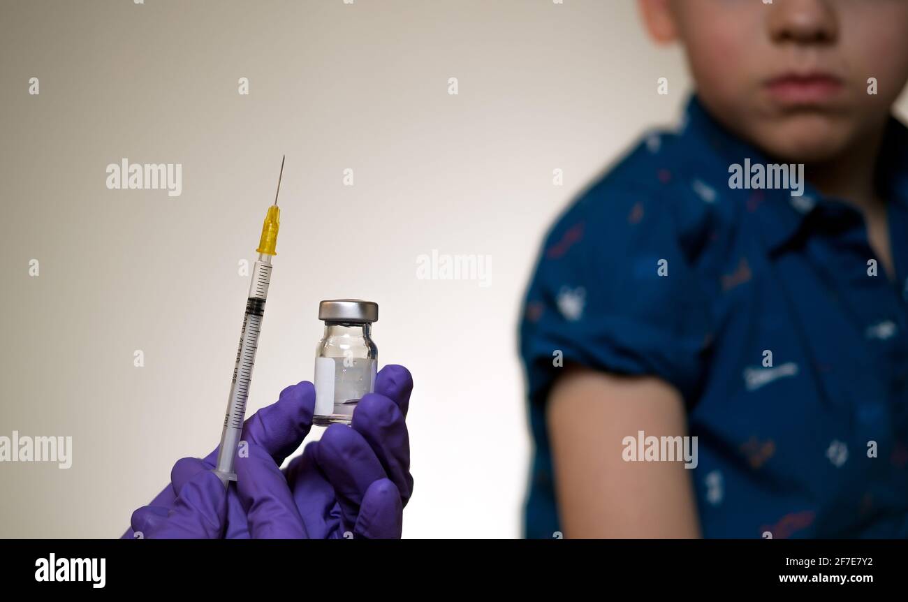 Vaccine vial hold in hand with syringe and child sitting with rolled up sleeve on the blurred background. Concept for children vaccination. Stock Photo