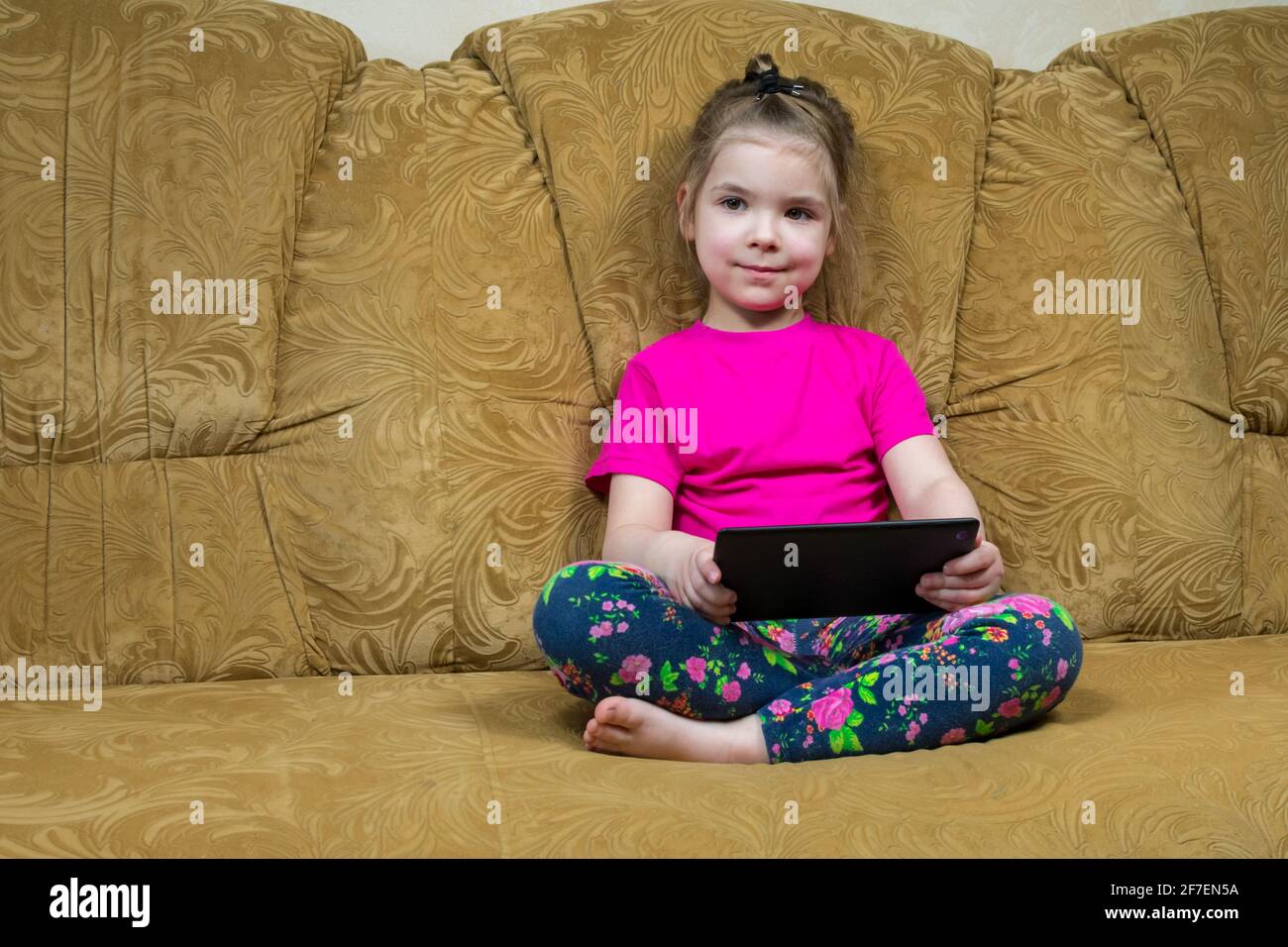Little girl sitting on a comfortable couch enjoying an online game on a digital tablet computer. Addicted to technology, happy little kid using fun Stock Photo