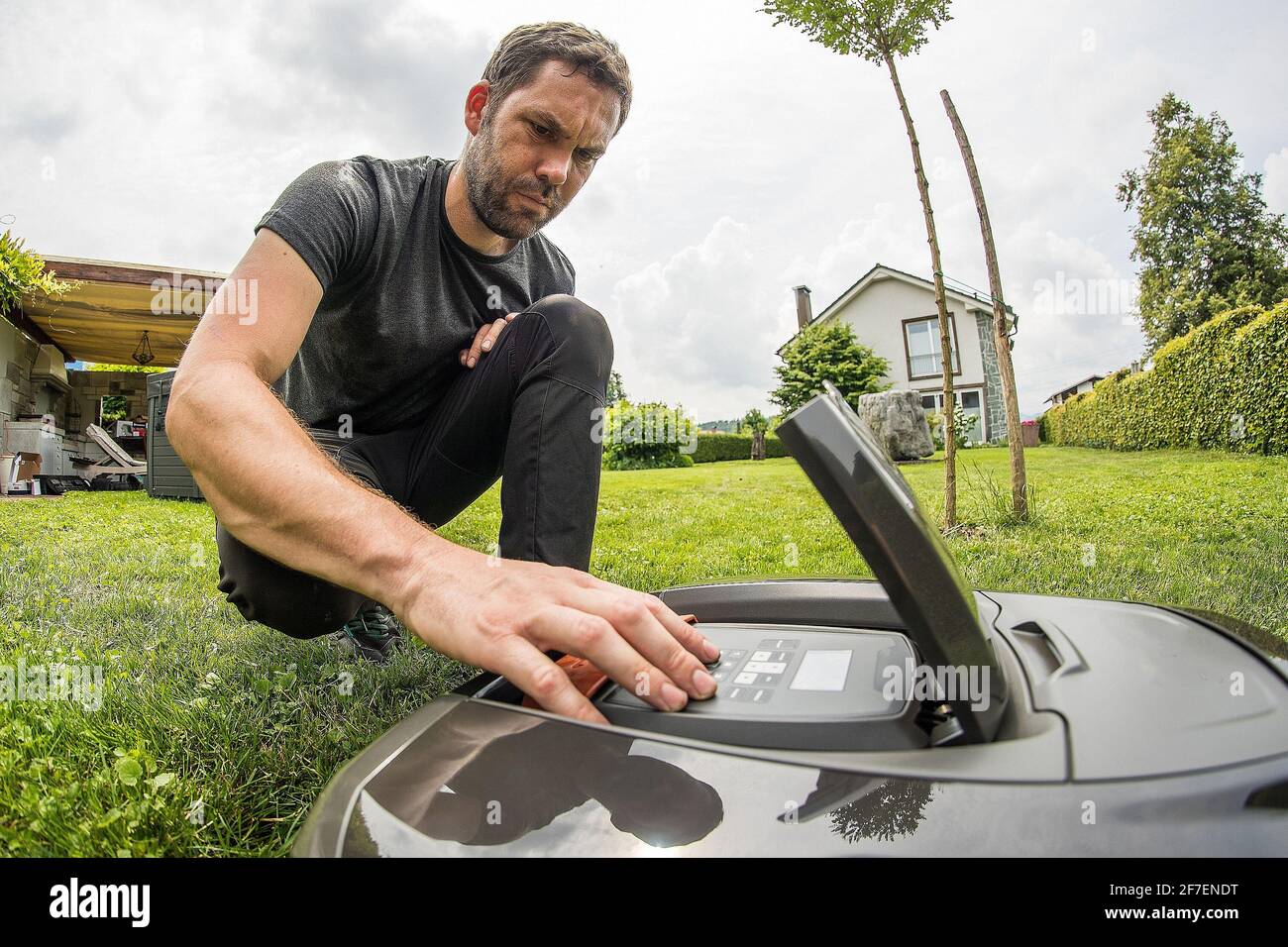 Young caucasian male is setting a robotic mower while kneeling in a nice green garden with blue skies. Stock Photo