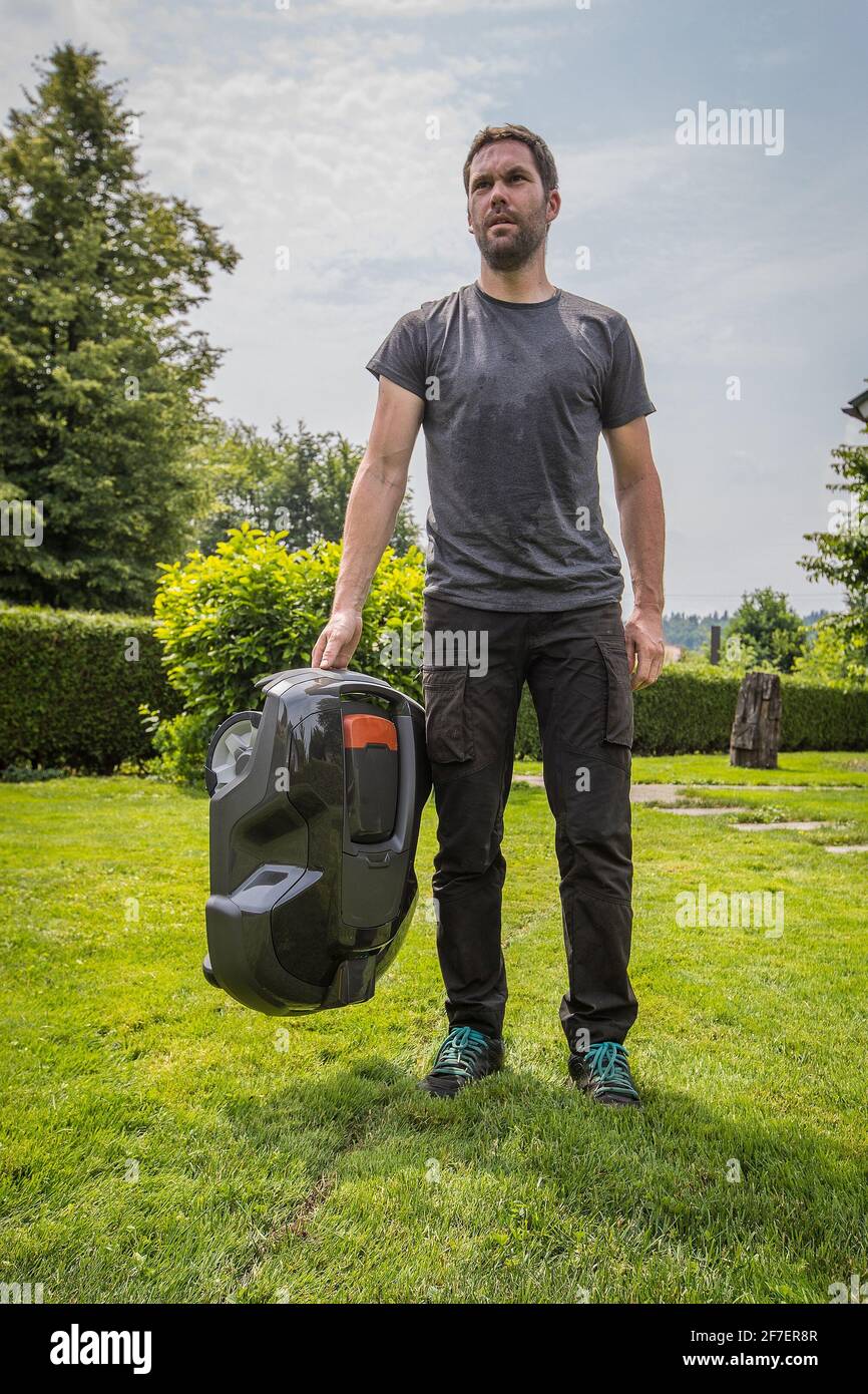 Young caucasian male is holding a robotic mower in his hand while standing in a nice green garden with blue skies. Stock Photo