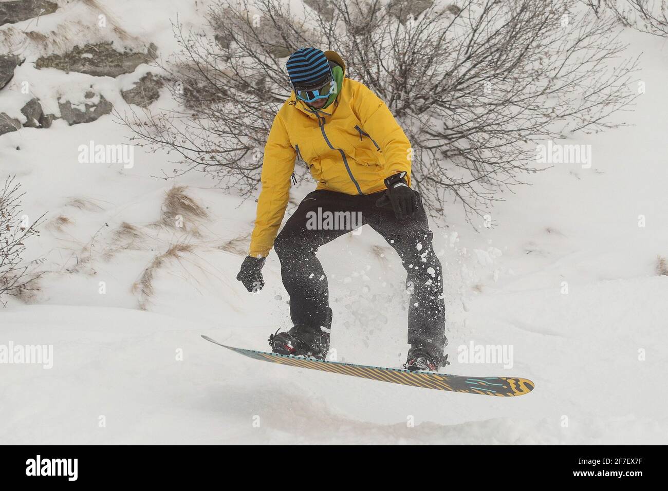 A snowboarder in snow going downhill amd jumping. Boarder with yellow clothing with black trousers and orange board riding and jumping over snow on a Stock Photo