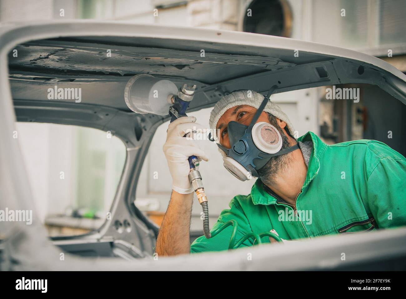 Caucasian man is spraying color with a compressed air paint gun on the vintage car as a restoration project. Man wearing protective equipment such as Stock Photo