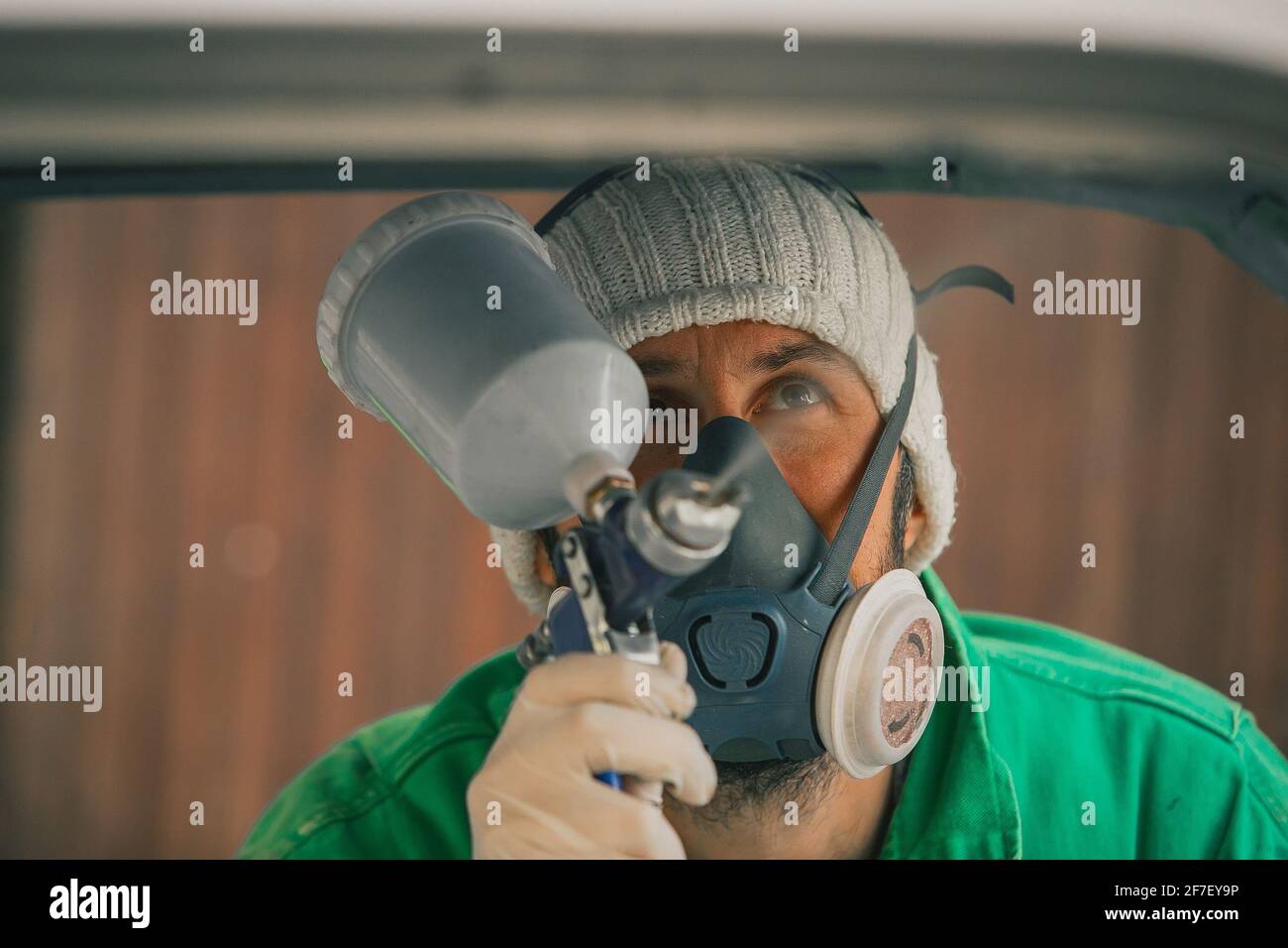 Caucasian man is spraying color with a compressed air paint gun on the vintage car as a restoration project. Man wearing protective equipment such as Stock Photo