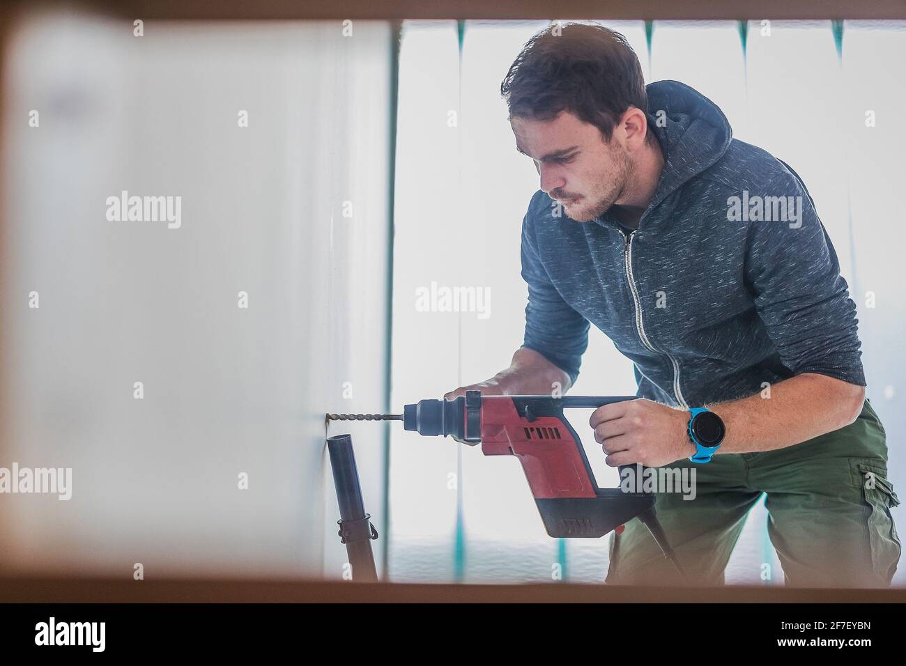 Man using an electric power drill to make a hole in the wall. Visible vacuum cleaner hose to suck in the debris. Stock Photo