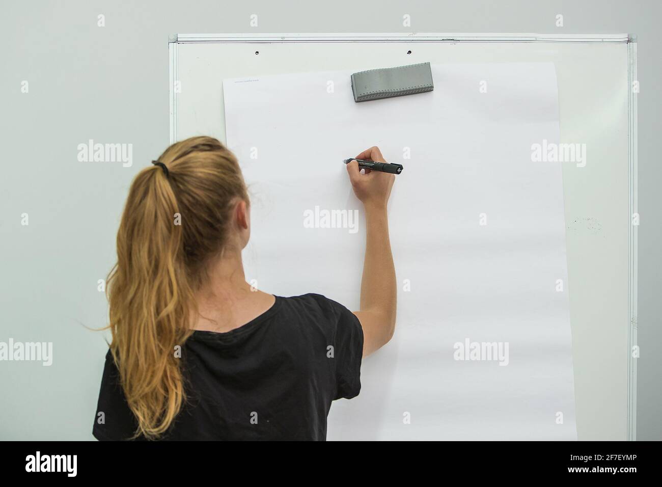 Young woman seen writing with a pen on a whiteboard or on paper. Focus on the pen, view from the back Stock Photo