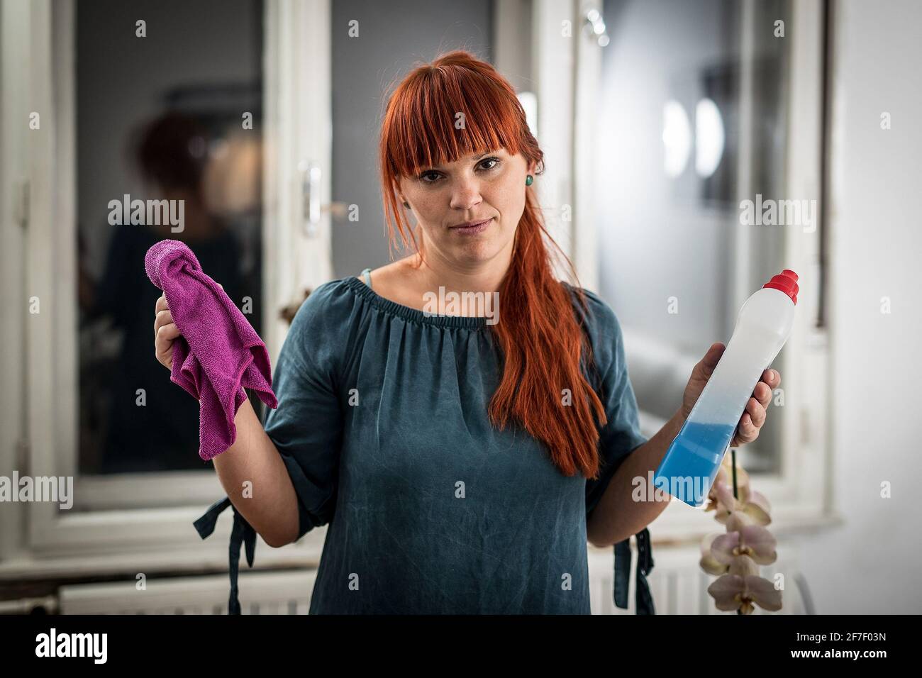 A young woman is sad and holding a bottle of cleaner and a mop in her hands, standing in a living room. Stock Photo