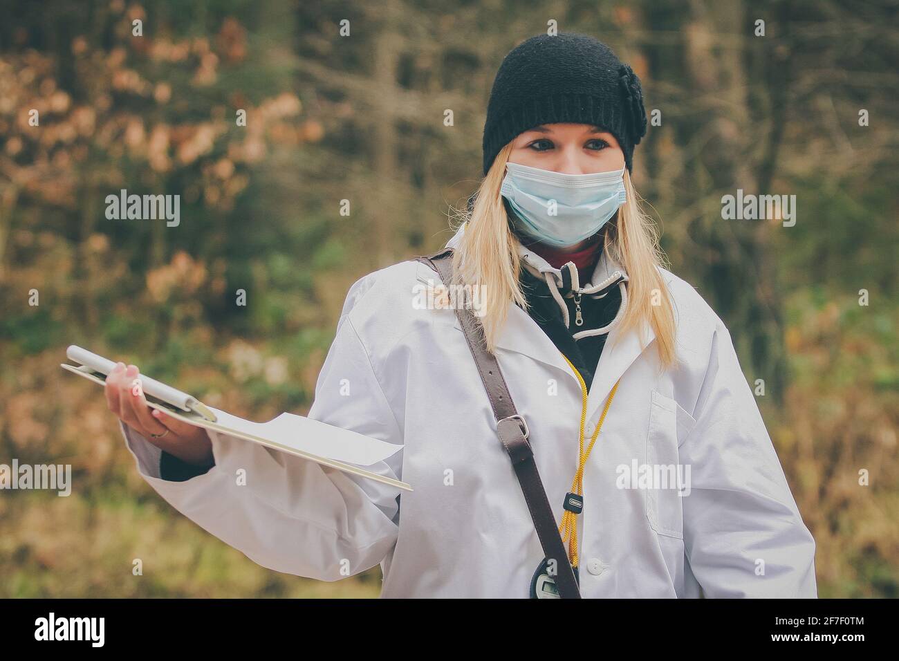 Young woman with blonde hair dressed as a doctor and a breathing face mask is standing outside in a cold autumn like forest. Stock Photo