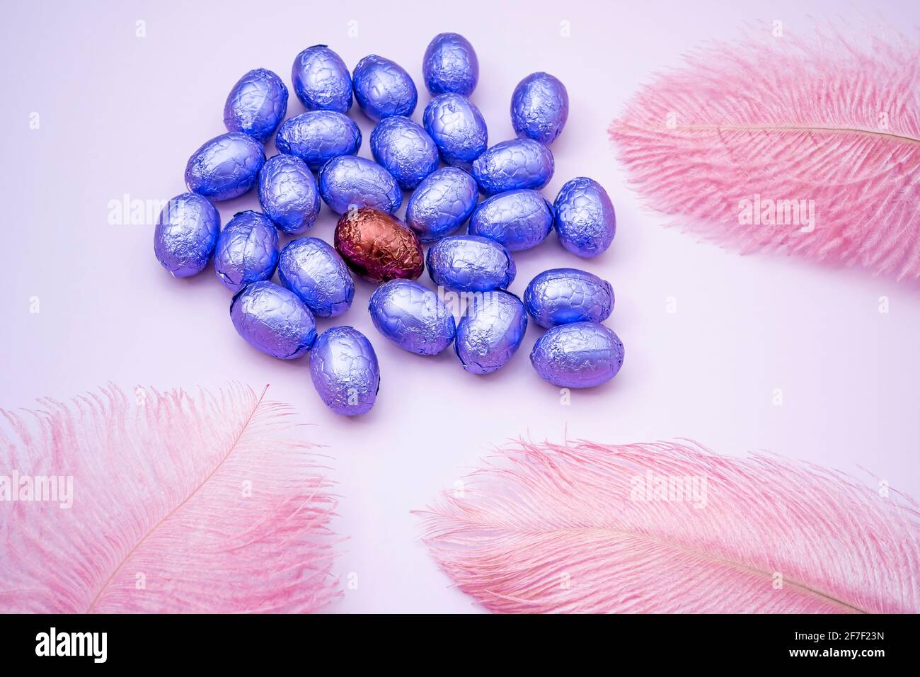 Easter pink background with chocolate easter eggs wrapped in purple colored foil with colorful feathers on a light rose background. Easter concept. Ae Stock Photo