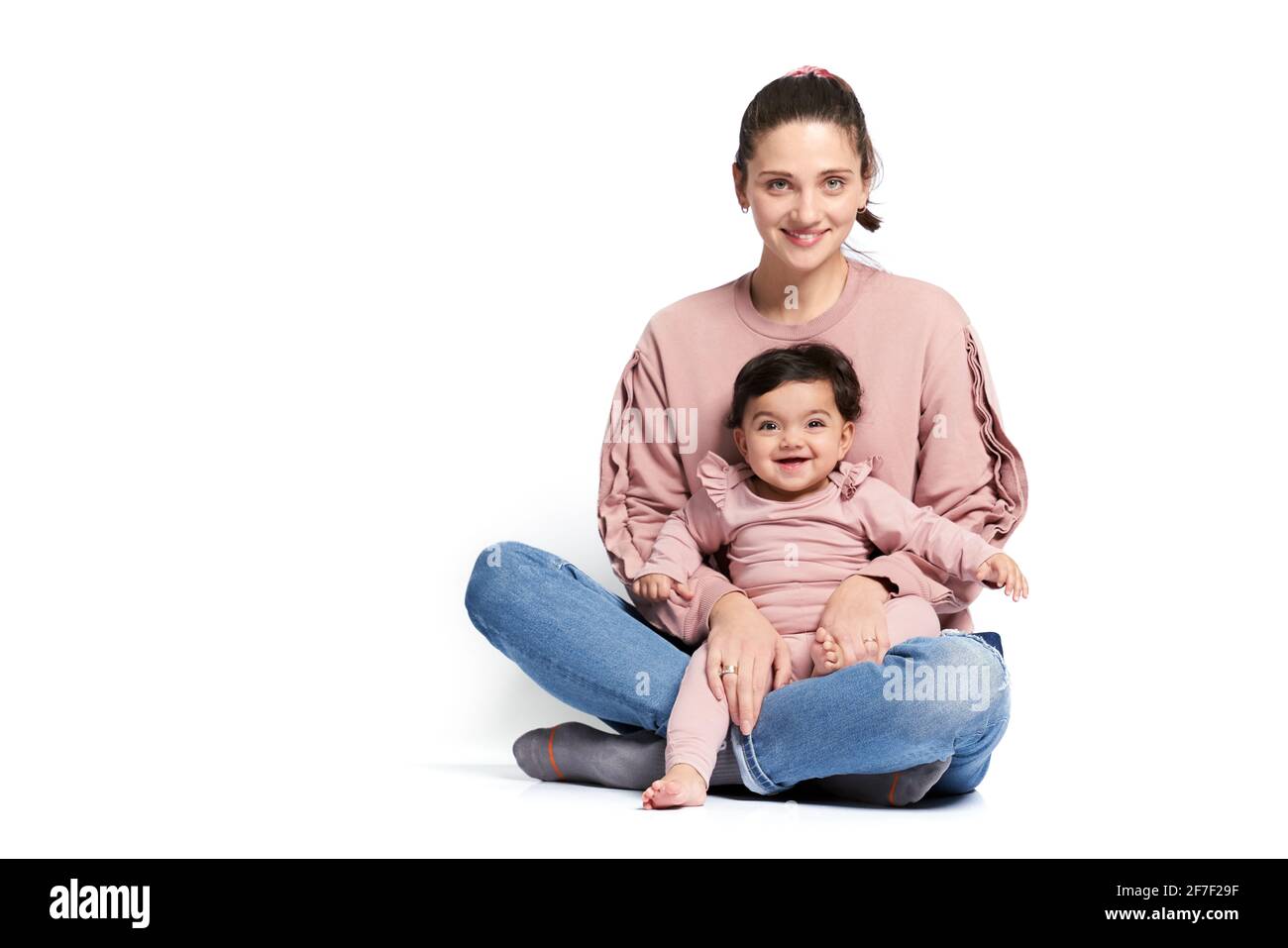 Portrait of cute mother with daughter looking at camera, isolated on white studio background. Young attractive woman holding sweet adorable child on leg while sitting on floor in lotus pose. Stock Photo