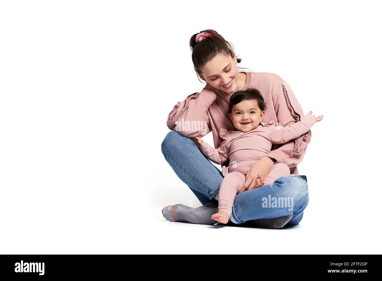 Portrait of cute mother with daughter posing isolated on white. Young woman holding sweet adorable baby girl on leg, looking at child and touching neck while sitting on floor in lotus pose. Stock Photo