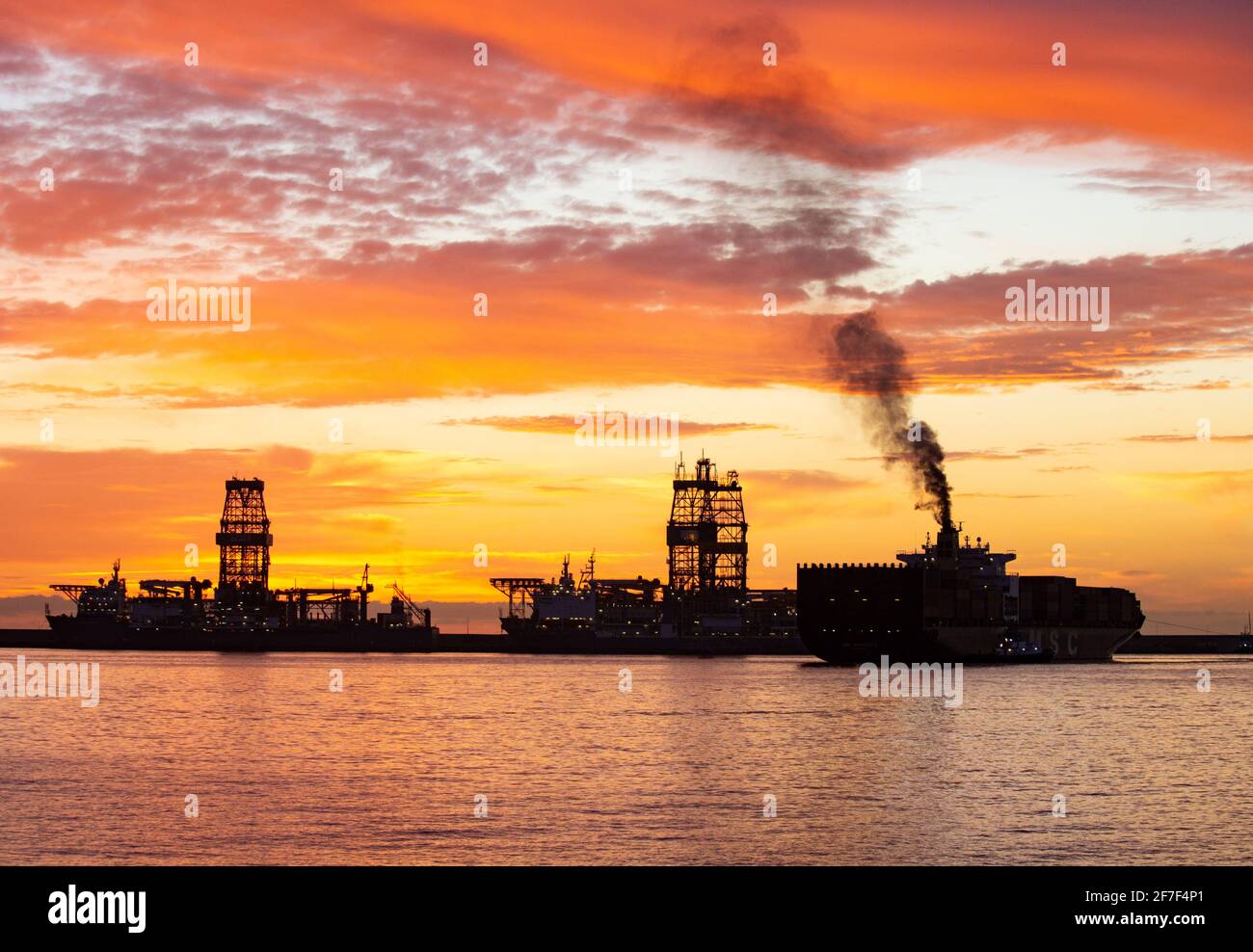 Las Palmas, Gran Canaria, Canary Islands, Spain. 7th April, 2021. A container ship belches out black smoke as the sky explodes with colour as the sun rises behind drilling ships in Las Palmas port on Gran Canaria. There are around ten drilling ships and oil rigs in the port which have been idle/mothballed for more than a year. Credit: Alan Dawson/Alamy Live News. Stock Photo
