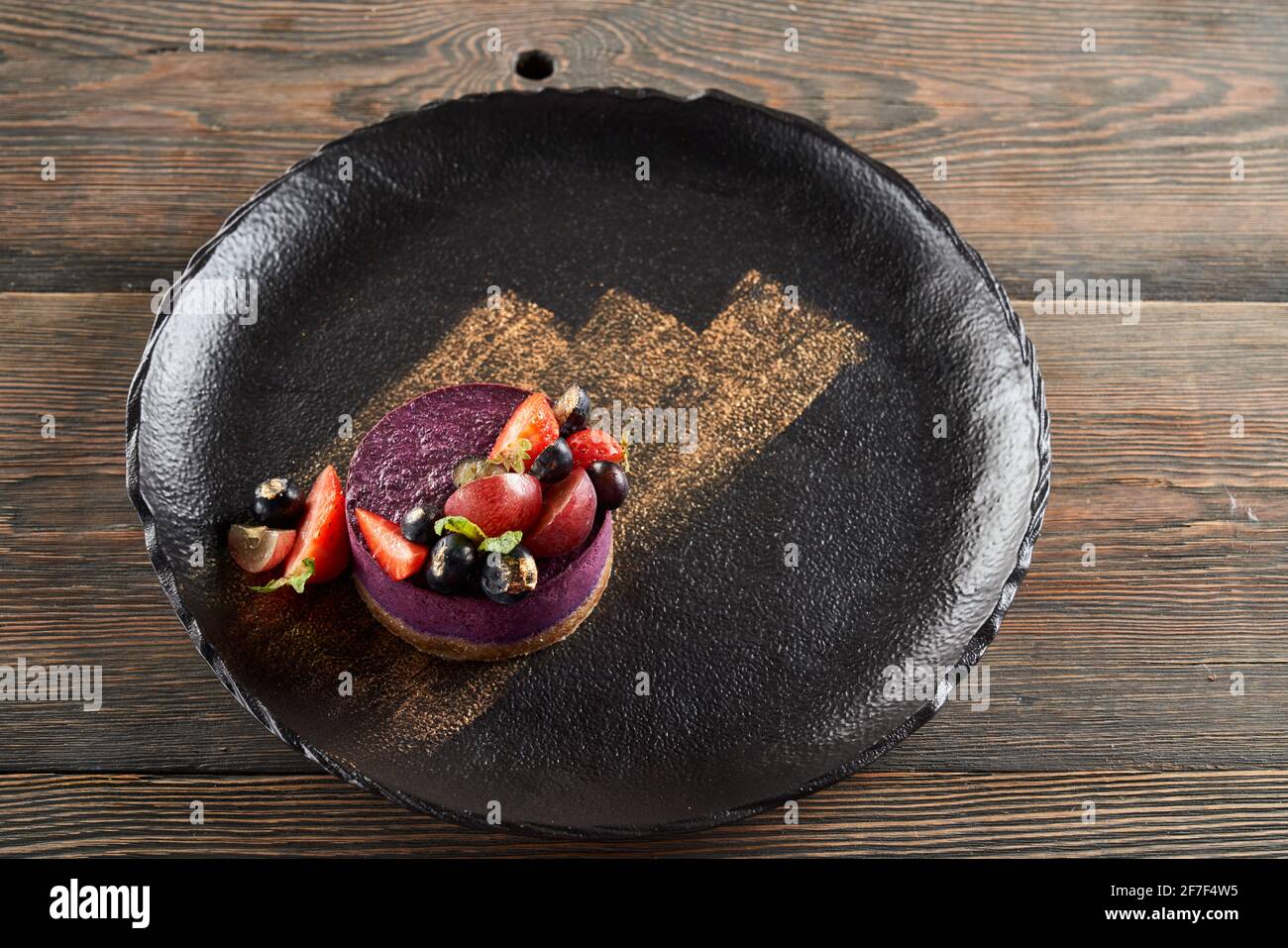 From above view of round piece of traditional blueberry cheesecake served with blueberries and strawberries on wooden table in restaurant. Golden brushstroke decoration on black plate. Stock Photo