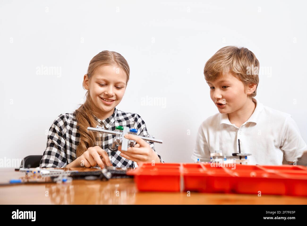 Interesting building kit for kids on table. Front view of boy and girl having fun, creating air vehicle. Science engineering. Nice interested friends smiling, chatting and working on project together. Stock Photo