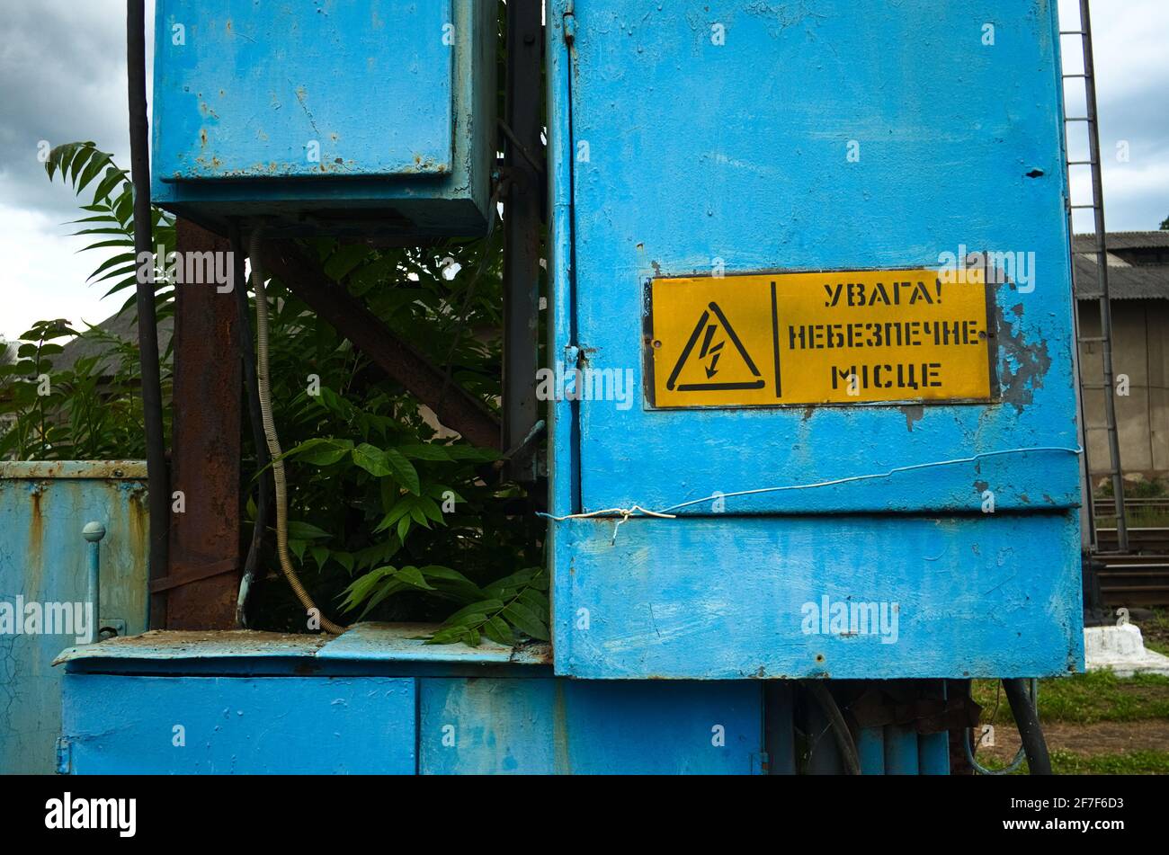 Old high voltage transformer sign on blue metal board. Electric power box near railroad tracks with danger triangle sign and caution notice on yellow Stock Photo