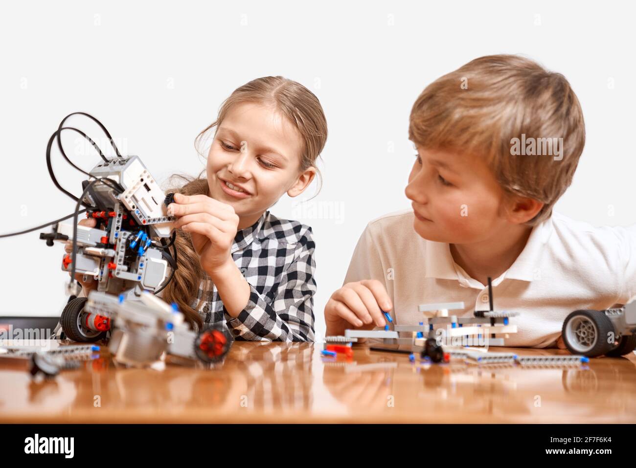 Front view of kids having fun, creating toys. Science engineering. Nice interested friends smiling, chatting, girl showing boy robot made with interesting building kit for kids on table. Stock Photo