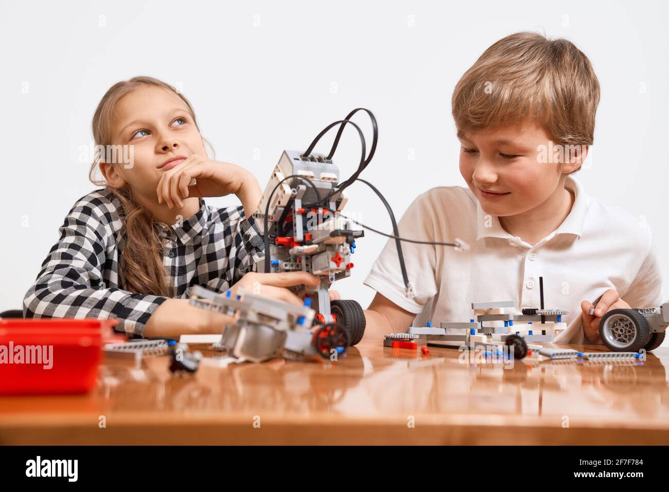 Building kit for kids on table. Front view of boy and girl having fun, creating vehicle. Science engineering. Nice interested friends working on project together, girl looking up and thinking. Stock Photo