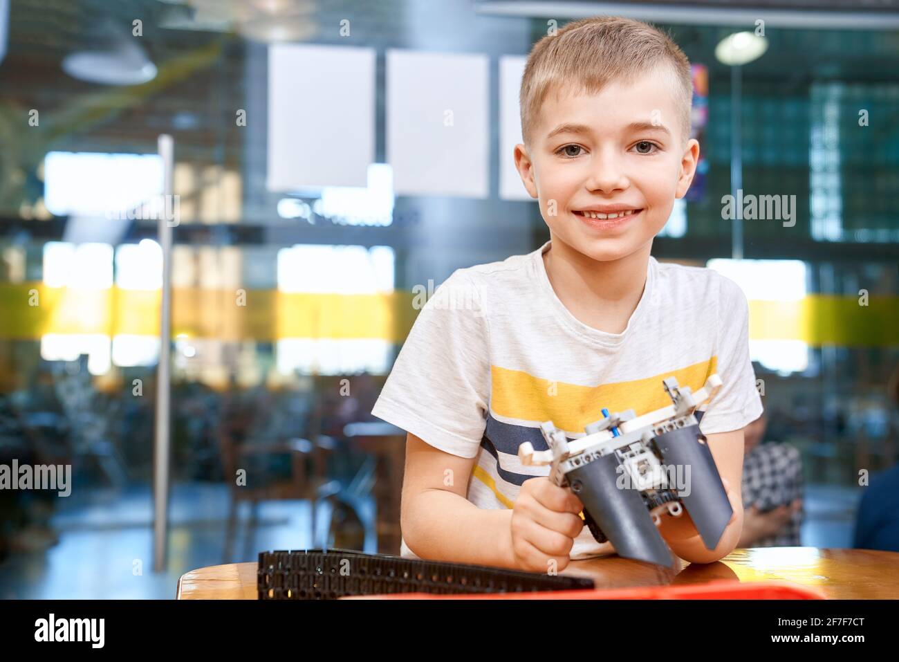 Front view of lovely caucasian boy smiling and looking at camera. Building kit for kids on table, children creating toys, having positive emotions and joy. Close up of boy working on project. Stock Photo