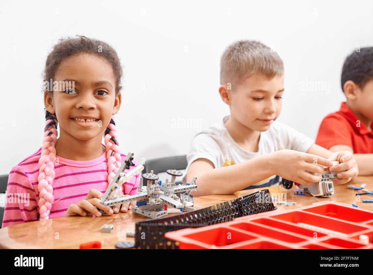 Side view of building kit for group of three multiracial kids creating toys, having positive emotions and joy, african girl smiling and looking at camera. Close up of friends working on project. Stock Photo