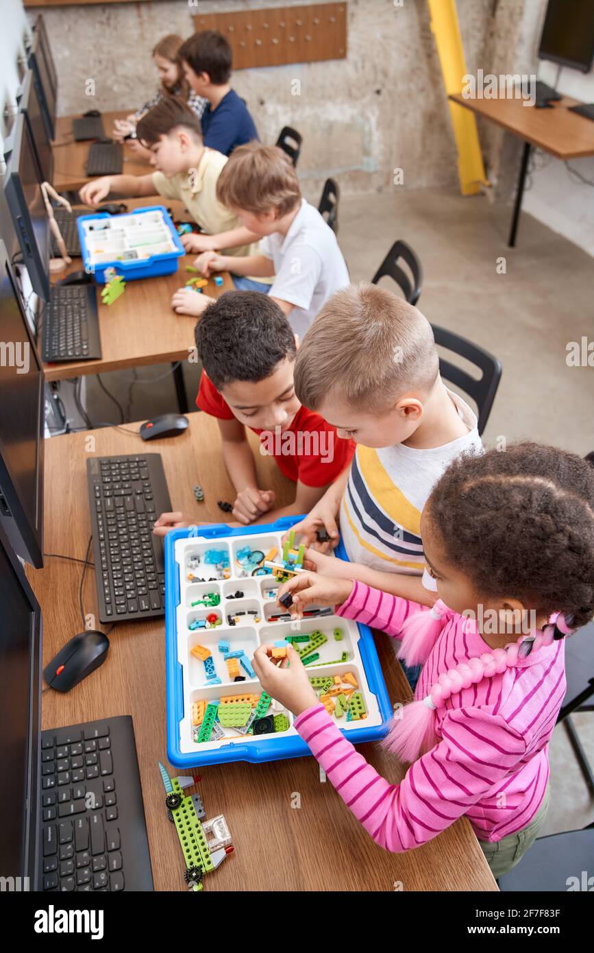Interesting building kit for kids on table with computers. From above view of boys and girls creating toys. Science engineering. Nice interested friends chatting and working on project together. Stock Photo