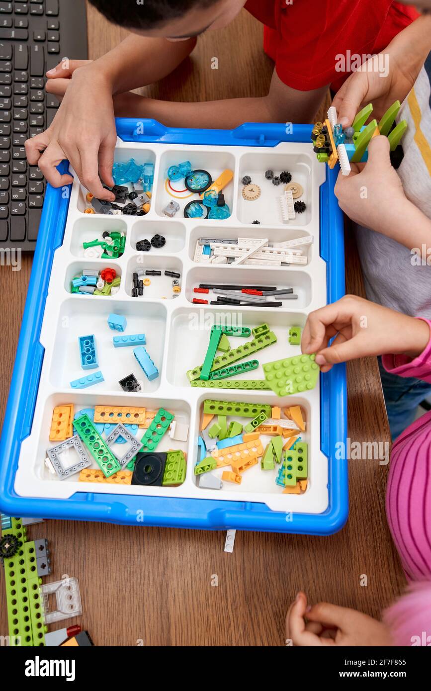 From above view of hands of incognito children taking details out of box and working on project. Colorful pieces from building kit for group of kids on table. Concept ofs cience engineering. Stock Photo